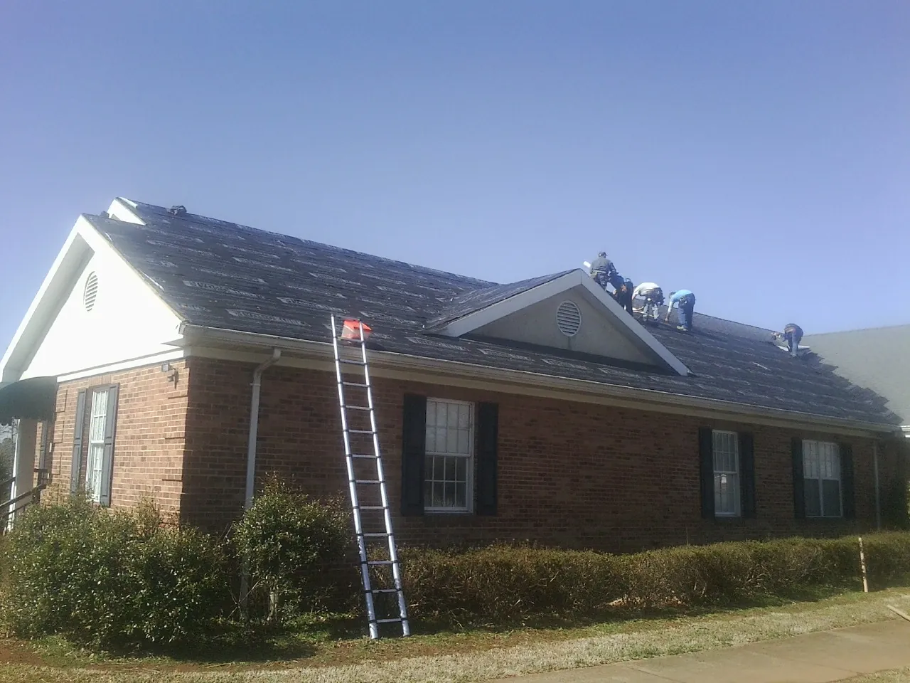 Crew tearing shingles off a brick ranch roof with a ladder set — Metal Roof Repair work by our crew in West Orange